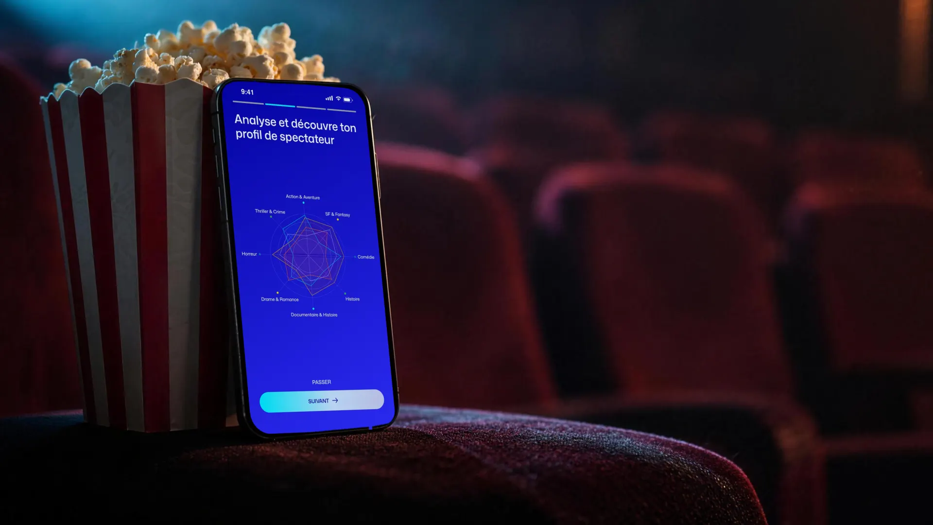 Un iPhone appuyé contre une boîte de pop-corn, sur l’accoudoir d’un fauteuil de cinéma. À l’écran, Klaps&nbsp;: «&nbsp;Analyse et découvre ton profil de spectateur&nbsp;» et le radar des genres.